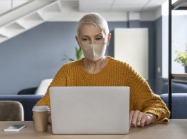 Foto de uma mulher idosa, de cabelos curtos, usando máscara, enquanto usa um laptop