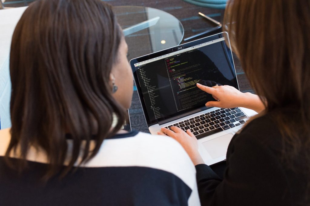 Foto de duas mulheres negras sentadas lado a lado, e uma delas aponta com o dedo indicador da mão direita para a tela de um laptop com vários códigos de programação