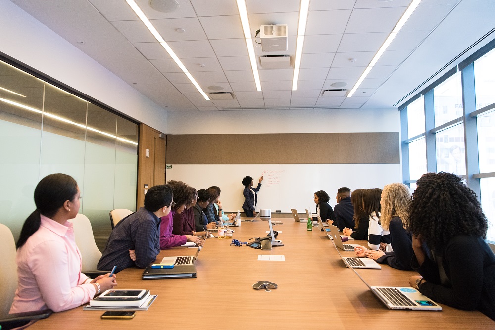 Foto de um grupo de pessoas de diferentes etnias e idades em uma sala, todos sentados, olhando em direção a uma mulher que está em pé e faz uma apresentação.