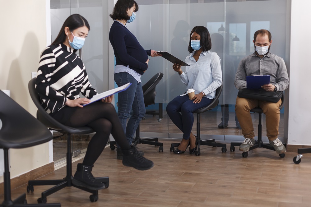 Foto de duas mulheres e um homem sentados em uma sala, enquanto uma mulher, em pé, entrega para outra uma prancheta. Todos usam máscara de proteção cobrindo a boca e o nariz.