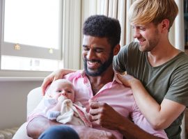 Foto de um casal de pais gays sorrindo para a filha bebê que está no colo de um deles, que segura a mãozinha esquerda da criança enquanto ela coloca a mão direita na boca.
