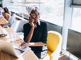 Foto de uma mulher negra sentada em uma cafeteria. Ela fala ao telefone enquanto a mão esquerda está em cima do mousepad do laptop.