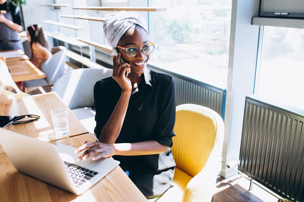 Foto de uma mulher negra sentada em uma cafeteria. Ela fala ao telefone enquanto a mão esquerda está em cima do mousepad do laptop.